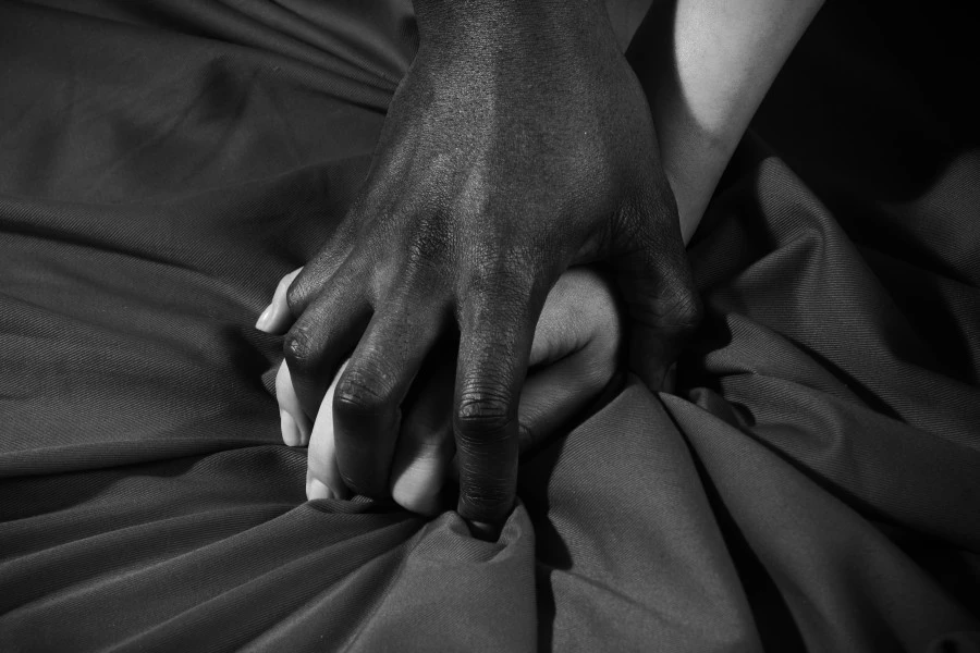 A man's hand over a women's hand that is clenching bed sheet.